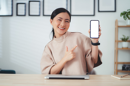 Asian woman showing smartphone with white screen mockup, sitting at home office, happy smiling and looking at camera.