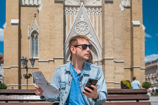 Man tourist sits on a bench in the city square with map and mobile phone and looks for hotel he booked from online application on his phone Male traveler looking for last minute booking. Travel, summer vacation concept