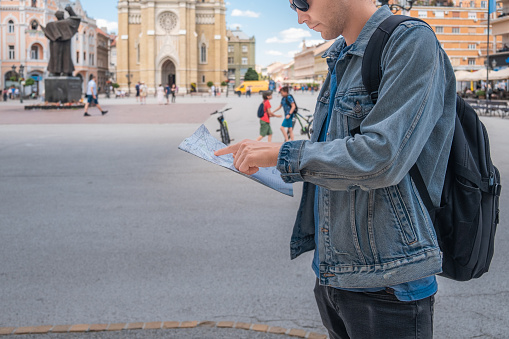 Man tourist on the square in the town with paper map looking is exploring new city. Handsome male traveler backpacker in sunglasses holding map and looking for attractions. Travel, summer vacation concept