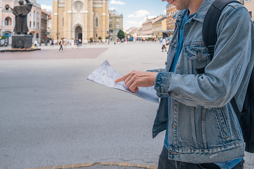 Man tourist on the square in the town with paper map looking for hotel he booked. Male traveler backpacker holding map and looking for attractions. Travel, summer vacation concept
