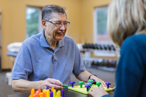 Cheerful senior man of Asian and Pacific Islander descent practicing fine motor skills with occupational hand therapist's assistance while at outpatient appointment at medical clinic.