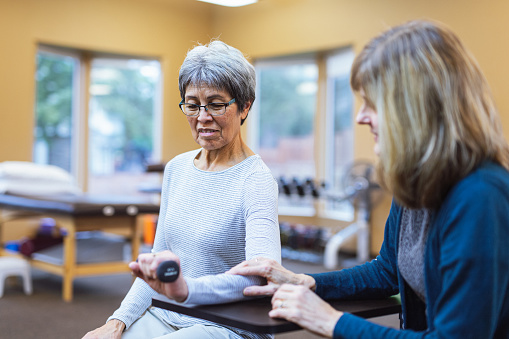 A senior woman of Pacific Islander descent does exercises with a small hand weight to build strength in her injured hand and wrist with the guidance of her female occupational therapist during an appointment at an outpatient rehabilitation clinic.