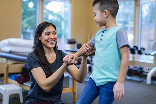 A multiracial female physical therapist smiles while providing care for a young Eurasian boy in the gym at an outpatient rehabilitation clinic.