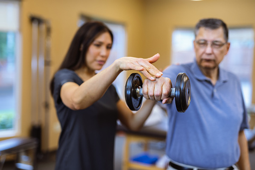 Selective focus on the hand of a senior man holding a dumbbell as his physical therapist provides guidance and support during a rehabilitation appointment at a medical clinic.