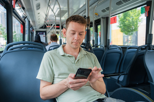 Attractive man is reading emails on his smartphone connected to public wi-fi while sitting in a city bus. Handsome male is looking at the screen of a mobile phone while texting message in social media