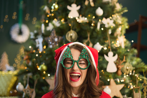 Christmas time. cheerful trendy woman with Santa hat and funny Christmas tree glasses near Christmas tree in the modern living room.