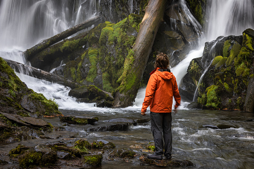 Hiker wearing an orange jacket standing in front of a large and beautiful waterfall in the Oregon forest