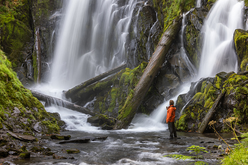 Hiker wearing an orange jacket standing in front of a large and beautiful waterfall in the Oregon forest