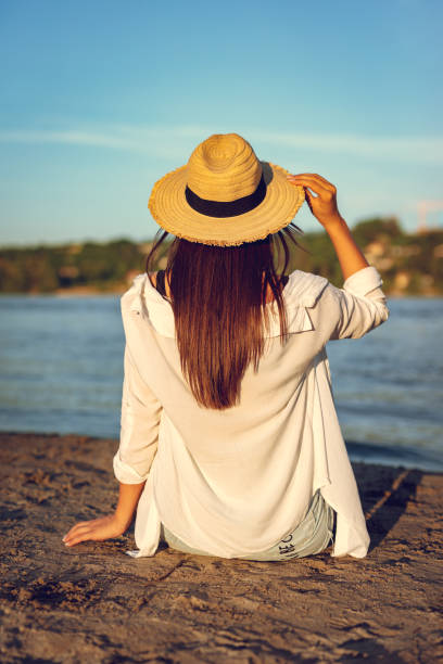 Rear view on a young woman in straw hat relaxing at sunset time on the river bank.