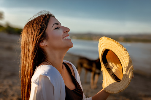 Portrait of a young smiling woman with a straw hat relaxing at sunset time on the river bank.