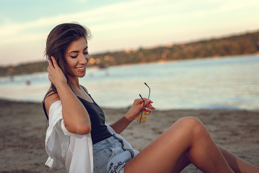 Young woman relaxing at sunset time on the river bank. She is sitting by the river and holding sunglasses.