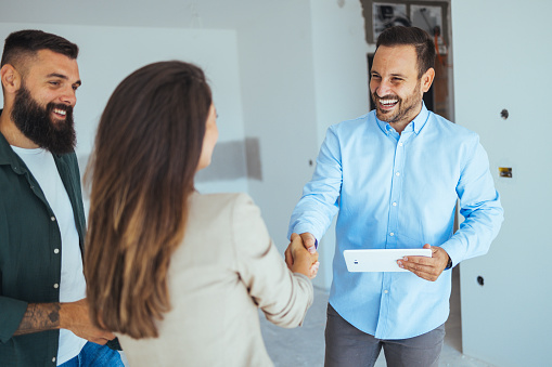 Real Estate Agent showing a house under construction to a couple and greeting them with a handshake at a construction site. Estate Agent Greeting Couple During Visit To New Home