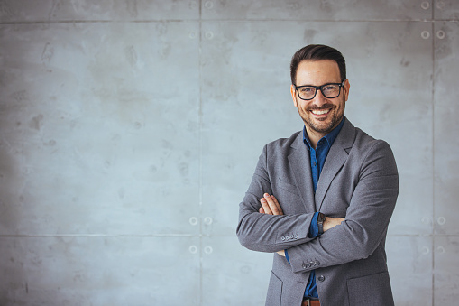 Portrait of happy businessman with arms crossed standing in office. Portrait of young happy businessman wearing grey suit and blue shirt standing in his office and smiling with arms crossed