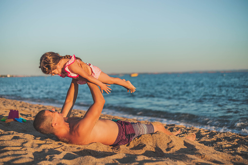 The man and his daughter are having a great time on the beach. The father is lying on the sand by the sea, and holding his little girl. The child is happily playing with her dad.