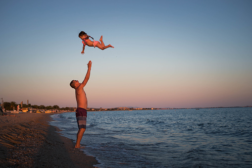 A man throws his daughter high and catches her while standing on the seashore. The girl is happy, because she can \