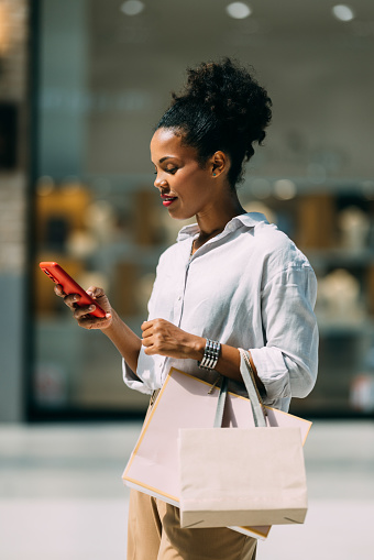 A smiling Latin female with shopping bags using her smartphone while standing on the street after going shopping.