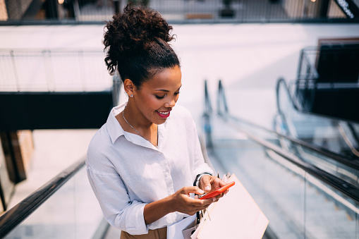 A smiling Latin female using her smartphone while standing on an escalator in a shopping mall.