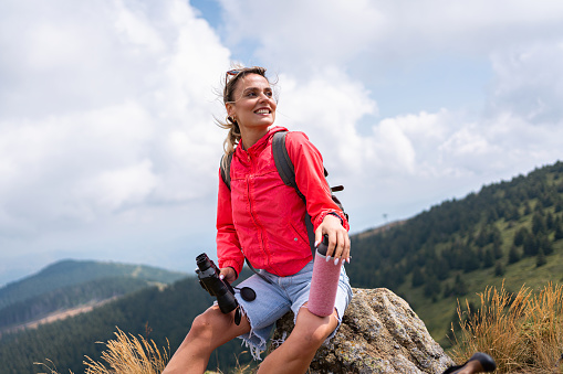 Active young Caucasian woman, sitting on a rock and holding binoculars, during her hike on the mountain