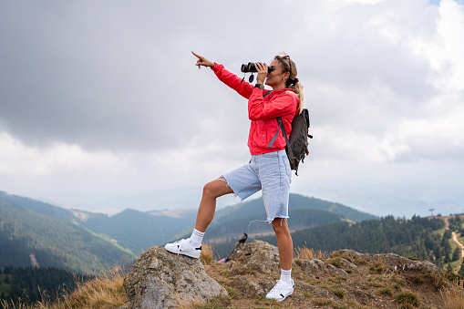 From the observation point on the mountain, a young Caucasian female hiker observing nature with binoculars