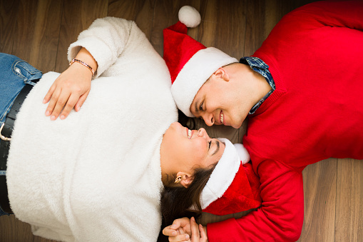 Romantic beautiful couple relaxing on the floor at home wearing santa hats ready to celebrate the christmas holidays