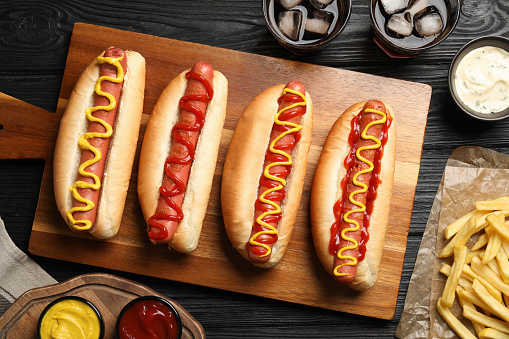 Delicious hot dogs with mustard, ketchup and potato fries on black wooden table, flat lay