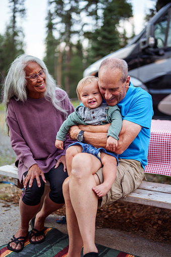 An adorable one year old Eurasian boy sits on his grandfather's lap and smiles directly at the camera as his adoring grandfather gives him a big hug. The multi-generational family is enjoying a fun summer camping trip.