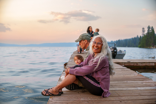 A beautiful and active senior woman of Hawaiian and Chinese descent sits on a dock with her multigenerational family and smiles directly at the camera while the sun sets over a lake in Oregon. The woman is on a fun and relaxing summer camping trip with her family.