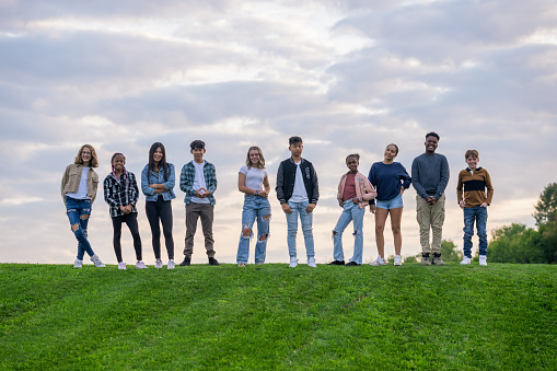 A large group of teens stand side-by-side at the top of a grassy hill as they pose together outside for a casual back-to-school portrait.  They are each dressed comfortably in fall layers and smiling.