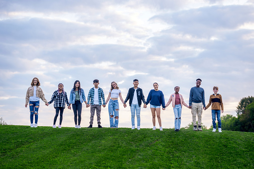A large group of teens stand side-by-side at the top of a grassy hill as they pose together outside for a casual back-to-school portrait.  They are each holding hands as they smile and enjoy the fresh air.