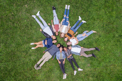 A large group of ethnically diverse teens, lay in the grass in a circle as they pose for a portrait.  They are each dressed casually and are smiling in this aerial view.