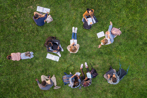 A large group of teens lay scattered in the grass outside as they individually study.  They are each dressed casually and are focused on their books in this aerial view.