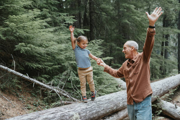 Joyful little girl enjoying nature with her playful grandfather An adorable Eurasian three year old girl who is balancing on top of a fallen log while hiking in a forest with her family holds hands with her active and playful grandfather as they both smile and cheer. grandparent stock pictures, royalty-free photos & images