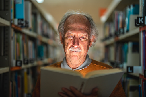 Portrait of a Bald senior Caucasian male with gray hair and mustaches reading a book. Standing between the bookshelves enjoying his book.