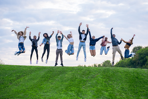 A large group of teens jump in the air at the top of a grassy hill as they celebrate going back-to-school.  They are each dressed in fall layers and smiling.