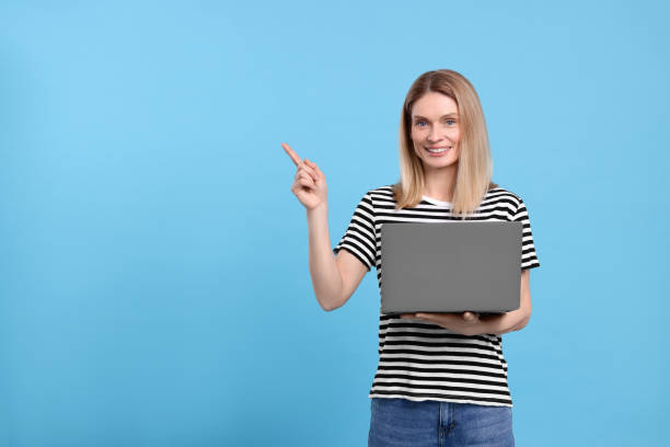 istock Happy woman with laptop pointing at something on light blue background. Space for text 1694603526