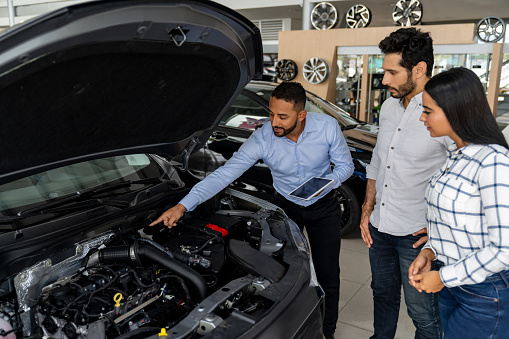 Car salesperson showing the engine of a car of a Latin American couple at the dealership - car ownership concepts