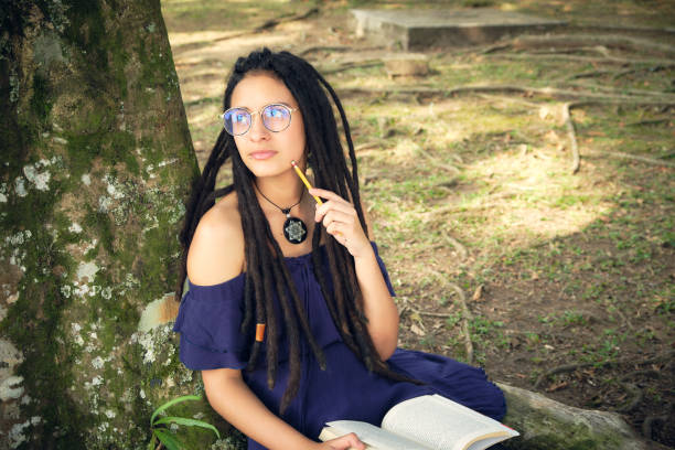 istock Girl with dreadlocks sitting under the tree, she is thinking and holding a book. 1694728481