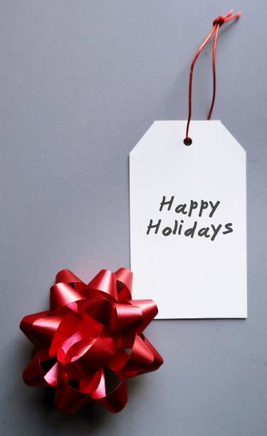 istock Red gift bow and white gift tag with hand written text HAPPY HOLIDAYS - all on gray background , concept of new year holiday gift for love ones 1694730781