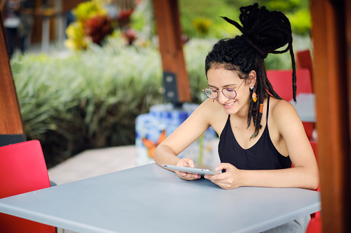 Young happy woman with dreadlocks reading from her tablet while sitting at a table, smiling and thinking about something positive.