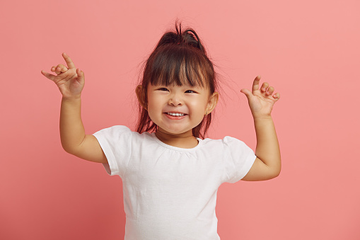 Joyful three years old Asian ethnicity little girl raises her hands up, expresses a cheerful mood, smiles happily wearing in white t-shirt standing on a pink isolated.