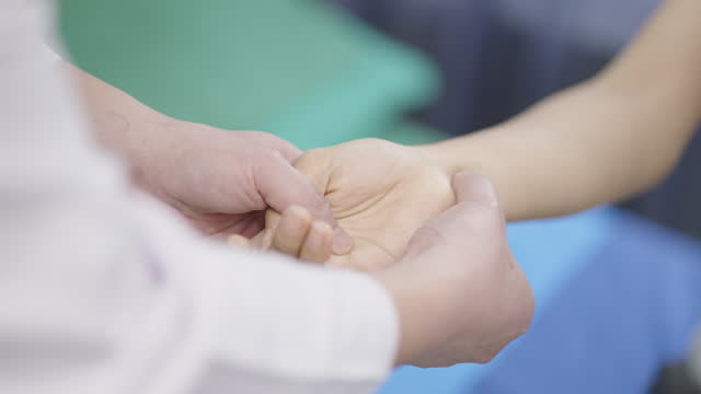 close-up of a patient who's having a wrist pain, sitting on an examination bed and stretching her arm to the doctor and letting him press each spot on her wrist to diagnose the problem at a medical clinic.