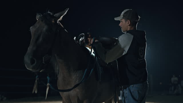 SLO MO Young Male Rancher Fastens Leather Saddle with Straps to Horse on Ranch at Night