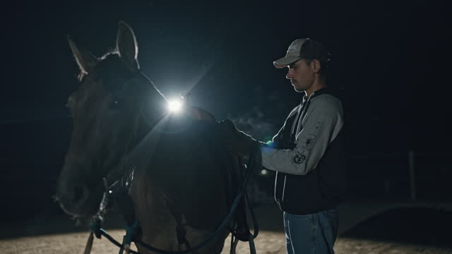 Young Male Rancher Dons and Secures a Leather Saddle onto his Horse on Serene Ranch at Night