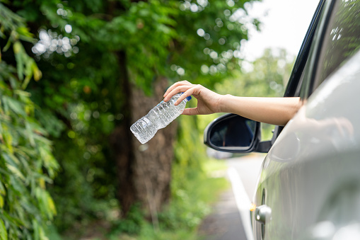 Woman's hand throwing away plastic bottle from car window on the road in green nature