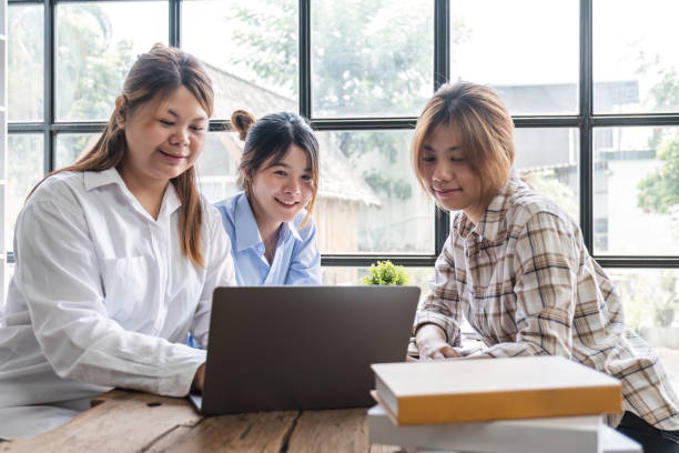 istock Asian College groups of students using laptop, tablet, studying together with notebooks documents paper for report near windows in classroom. Happy young study for school assignment, Soft focus 1694872474