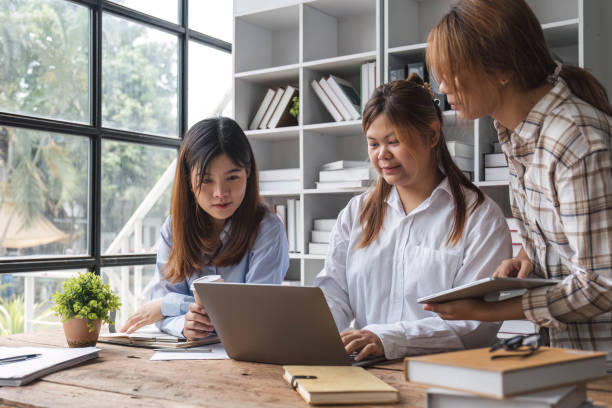 istock Asian College groups of students using laptop, tablet, studying together with notebooks documents paper for report near windows in classroom. Happy young study for school assignment, Soft focus 1694873554
