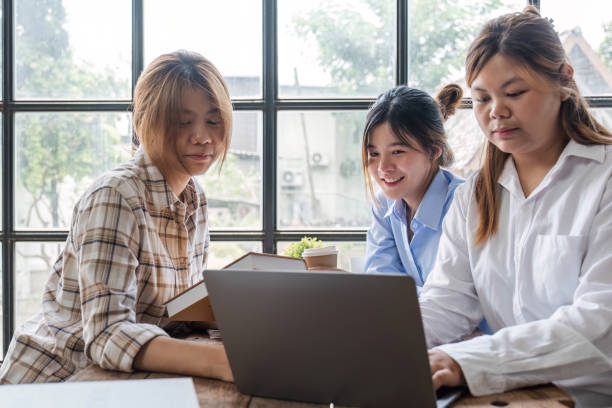 istock Asian College groups of students using laptop, tablet, studying together with notebooks documents paper for report near windows in classroom. Happy young study for school assignment, Soft focus 1694876434