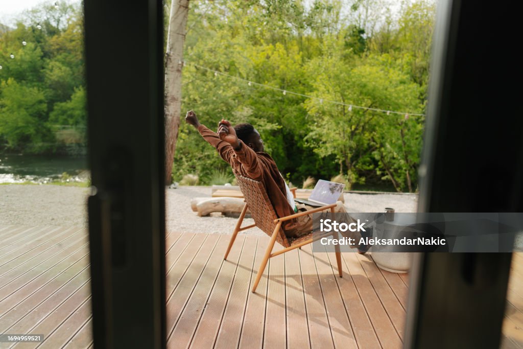Working remotely from the log cabin on the riverbank Photo of a young African American man, working from a remote location on his  computer Rear View Stock Photo