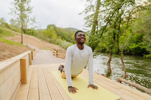 Photo of a young African American man working out early in the morning, right after he starts his day. He is working out outdoors on decking near to the river.