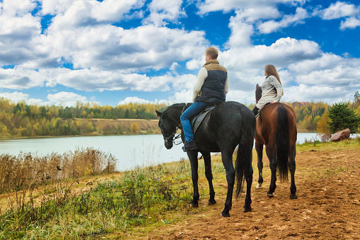 Rear view of couple man and woman on horses in fall forest with lake by blue sky. From behind riders on horseback in autumn park, rural scene. Leisure activity and resting concept. Copy ad text space